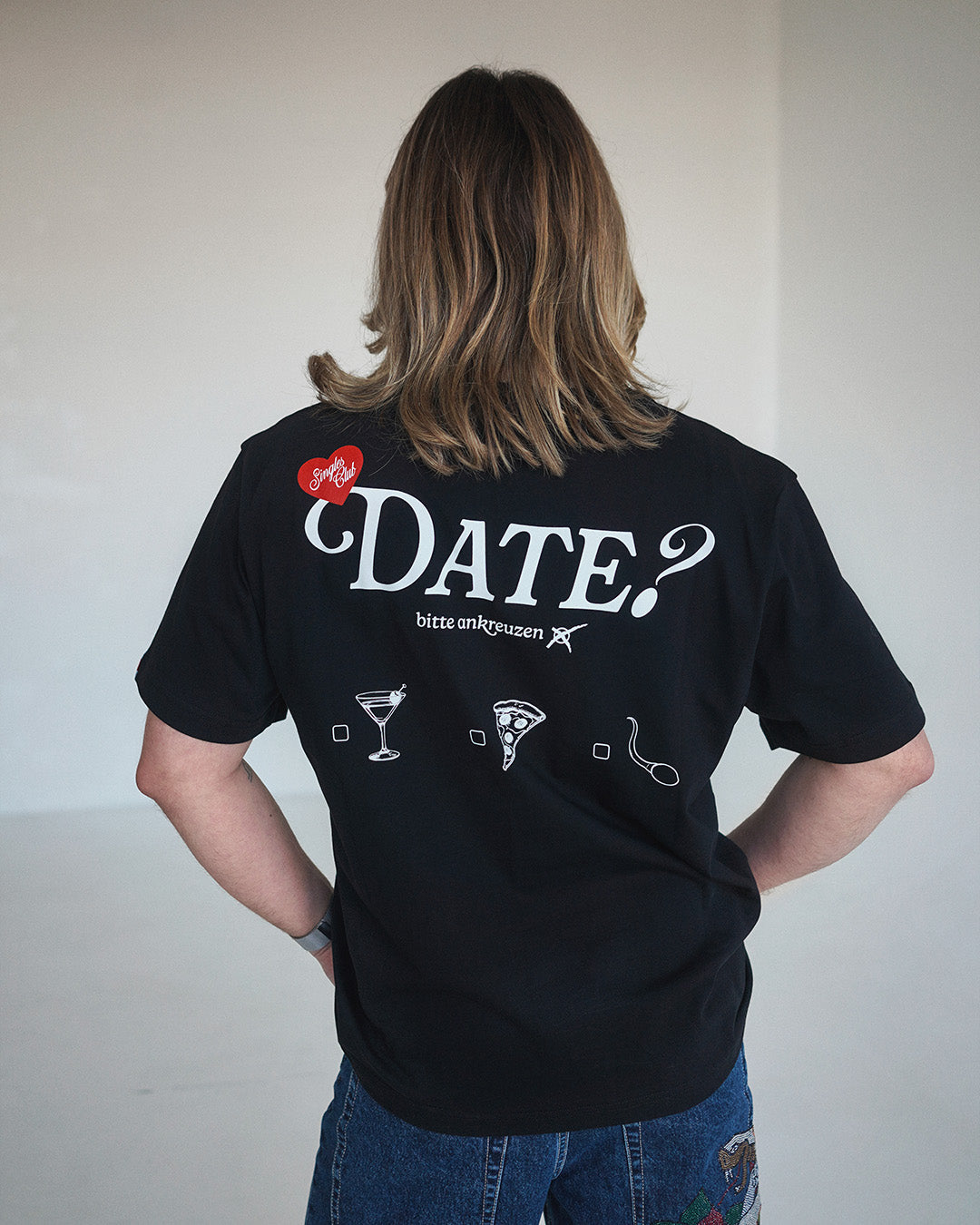 Rückansicht einer Person im schwarzen T-Shirt mit dem weißen Aufdruck „DATE? bitte ankreuzen“ und Auswahlmöglichkeiten: Cocktail, Pizza oder Sperma.