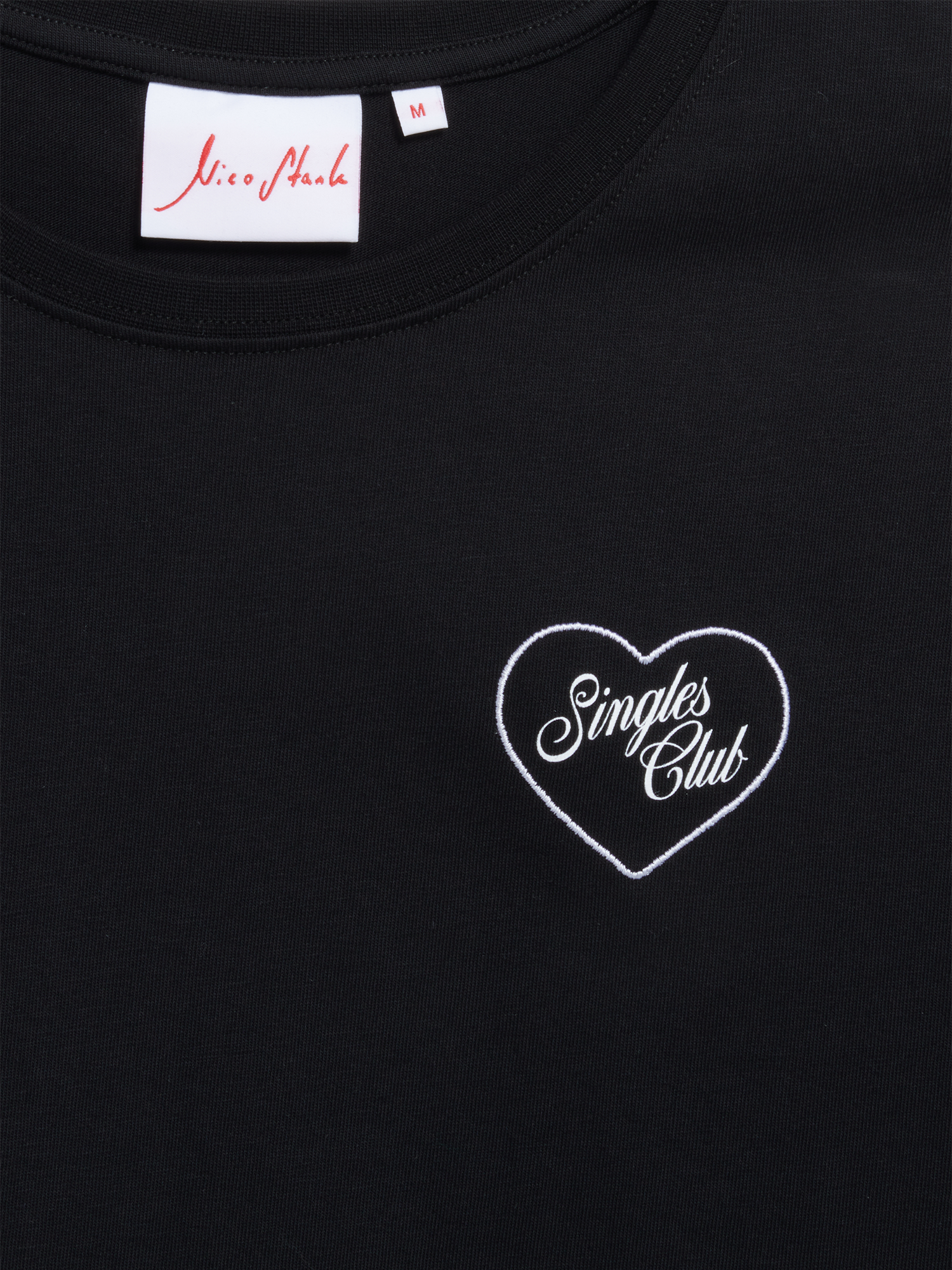Nahaufnahme des gestickten Herz-Logos mit „Singles Club“-Schriftzug auf schwarzem T-Shirt-Stoff.